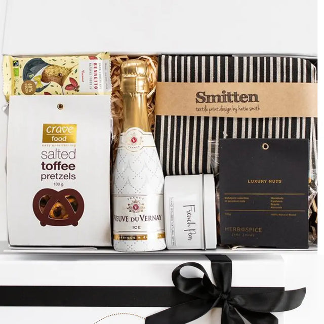 Gift basket with champagne, snacks, and a book inside a box with a black ribbon.