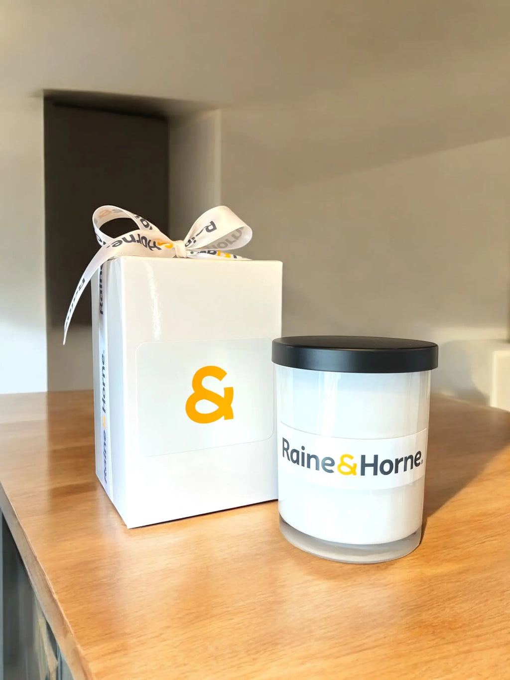 White gift box with a bow and a branded candle on a wooden surface.