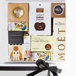 A luxury gift box containing a variety of gourmet treats including champagne, meringues, pretzels, chocolate bars, hand cream, and a candle, all beautifully packaged in a white box with tissue paper and a ribbon.