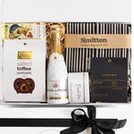 Gift basket with champagne, snacks, and a book inside a box with a black ribbon.