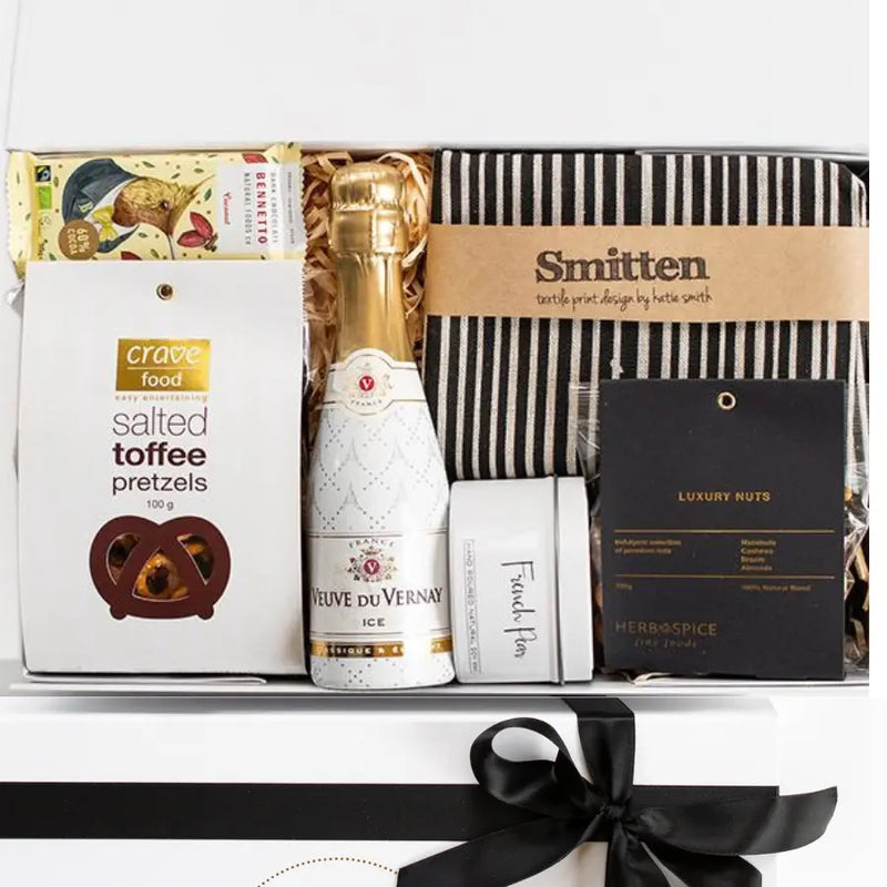Gift basket with champagne, snacks, and a book inside a box with a black ribbon.