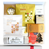 A gift box containing a variety of food items including gingerbread trees, salted caramel nut mix, butterscotch and brandy sauce, chocolate brownie bite, champagne bears in a Christmas cracker, vanilla meringues, maple and chili cashew nuts, and a handmade candle, all packaged in a white box with a gold seal and ribbon.