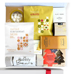 A gift box containing a variety of food items including gingerbread trees, salted caramel nut mix, butterscotch and brandy sauce, chocolate brownie bite, champagne bears in a Christmas cracker, vanilla meringues, maple and chili cashew nuts, and a handmade candle, all packaged in a white box with a gold seal and ribbon.