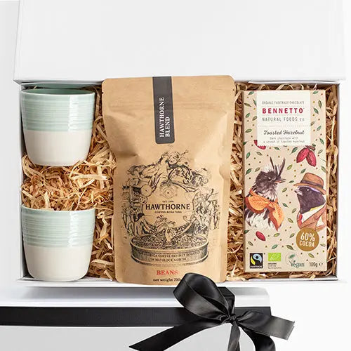 Gift set with coffee beans, cups, and a bar of chocolate in a box with a black ribbon.