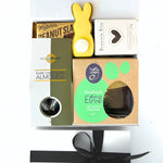 An assortment of chocolate themed gifts including a chocolate-dipped shortbread egg, chocolate fudge brownie bite, iced gingerbread Easter bunny cookie, and dark chocolate almonds, all packaged in a white gift box with a gold ribbon.