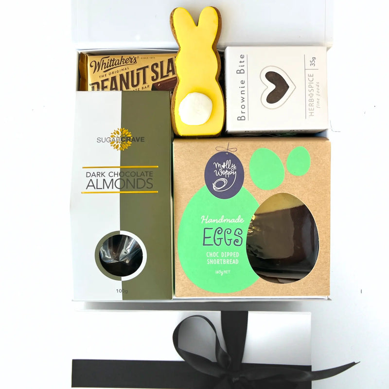 An assortment of chocolate themed gifts including a chocolate-dipped shortbread egg, chocolate fudge brownie bite, iced gingerbread Easter bunny cookie, and dark chocolate almonds, all packaged in a white gift box with a gold ribbon.