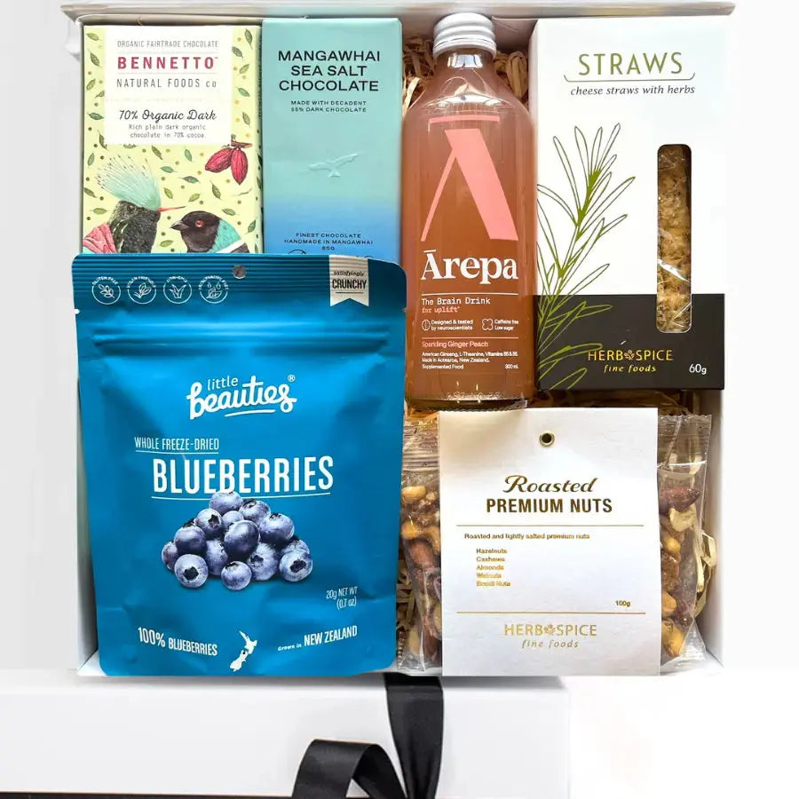 An assortment of gourmet food items in a gift box, including Bennetts dark chocolate bar, Mangawhai sea salt chocolate, Arepa sparkling ginger and peach drink, roasted premium nuts, blueberries, and Herb and Cheese straws.