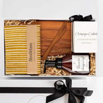 Gift set with a bottle, wooden spoon, and striped towel in a box with a black ribbon.