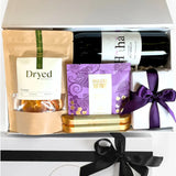 Gift basket with wine, snacks, and a book tied with a purple ribbon.