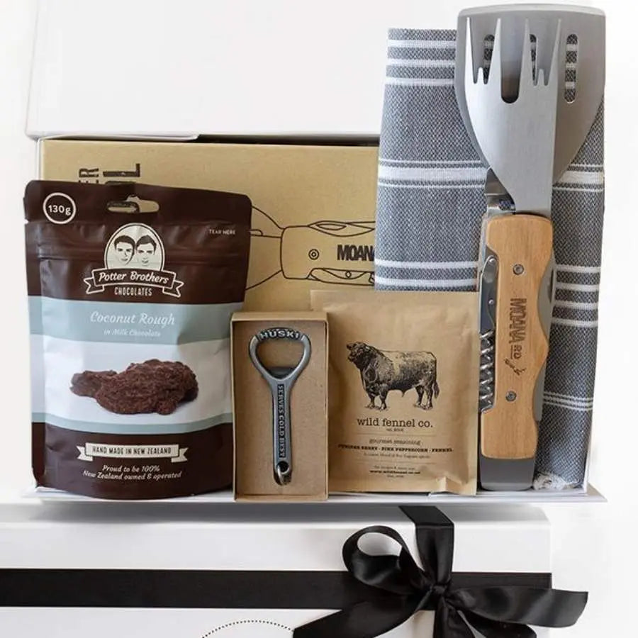 A gift box containing a variety of items including a BBQ tool set, a Turkish towel, a bottle opener, and a packet of chocolate chunks. The items are displayed neatly in a white box with a black ribbon.