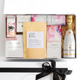 A luxury white gift box with magnetic closure containing a variety of pampering gifts including a bottle of champagne, chocolate with raspberry, bath bombs, a hand cream, a natural soy candle, and a keepsake gift card.