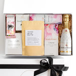A luxury white gift box with magnetic closure containing a variety of pampering gifts including a bottle of champagne, chocolate with raspberry, bath bombs, a hand cream, a natural soy candle, and a keepsake gift card.