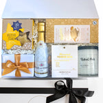 A luxury gift box with a variety of items including a bottle of champagne, a chocolate fudge brownie, a block of caramelised white chocolate, a premium nut mix, a buttery shortbread, a soy candle, and a keepsake box with a gold or red ribbon.