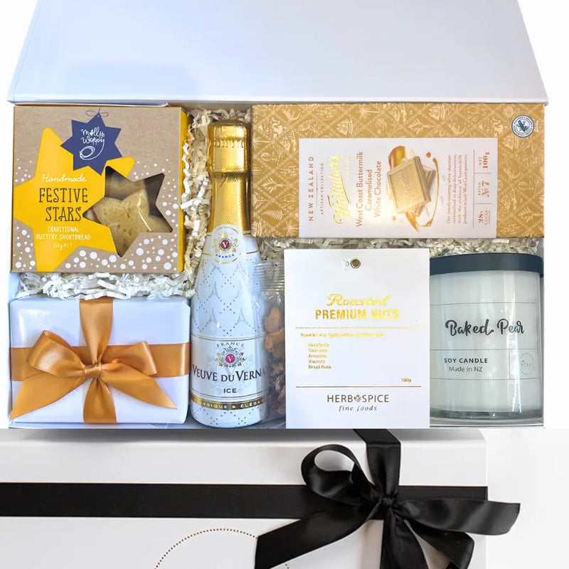 A luxury gift box with a variety of items including a bottle of champagne, a chocolate fudge brownie, a block of caramelised white chocolate, a premium nut mix, a buttery shortbread, a soy candle, and a keepsake box with a gold or red ribbon.