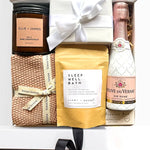 Gift set with a bottle of champagne, candle, bath salt package, and knitted blanket.