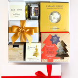 A luxury white gift box with magnetic closure containing a variety of Christmas treats, including a candle, caramel pebbles, butterscotch and brandy sauce, gingerbread Christmas trees, chocolate brownie bite, and fig relish, all decorated with a gold ribbon and a personal message card.