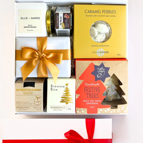 A luxury white gift box with magnetic closure containing a variety of Christmas treats, including a candle, caramel pebbles, butterscotch and brandy sauce, gingerbread Christmas trees, chocolate brownie bite, and fig relish, all decorated with a gold ribbon and a personal message card.