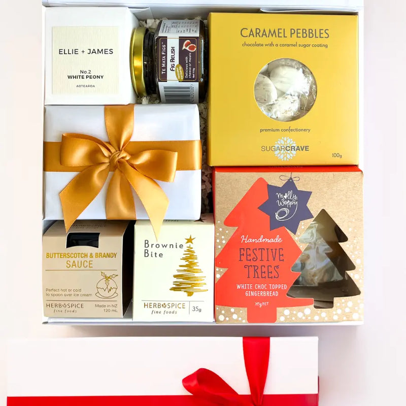 A luxury white gift box with magnetic closure containing a variety of Christmas treats, including a candle, caramel pebbles, butterscotch and brandy sauce, gingerbread Christmas trees, chocolate brownie bite, and fig relish, all decorated with a gold ribbon and a personal message card.