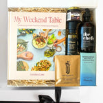 Gift box with 'My Weekend Table' book, bottles, and other items on a white background
