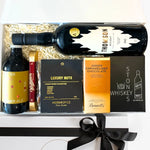 A gift box containing a bottle of Thomson whiskey, a bottle of ginger ale, a set of whiskey stones, a luxury nut mix, a caramelized chocolate bar, and a keepsake gift box with tissue paper and a black ribbon.