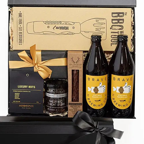 A gift box containing a variety of items including luxury nuts, a BBQ tool, organic chocolate, gourmet relish, and craft beer bottles.