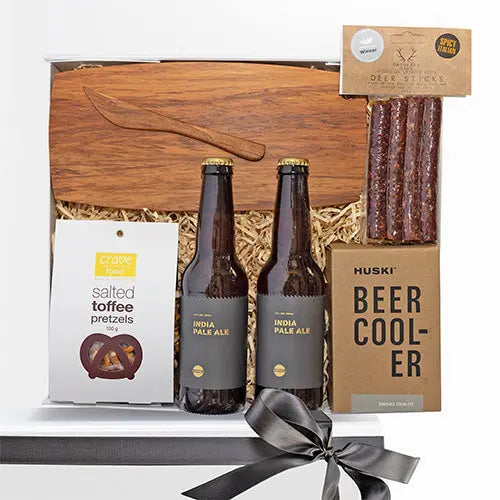 A gift box containing a cheese board, salted toffee pretzels, two bottles of India Pale Ale, a beer cooler, and deer sticks, all arranged with decorative elements like tissue paper and a keepsake card.