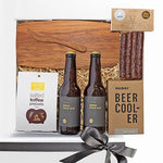 A gift box containing a cheese board, salted toffee pretzels, two bottles of India Pale Ale, a beer cooler, and deer sticks, all arranged with decorative elements like tissue paper and a keepsake card.