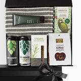 A luxury black gift box with magnetic closure containing a variety of gift items including a Turkish hand/gym towel, men's daily face wash, two cans of craft beer, a box of herb and cheese straws, and a salted toffee pretzel snack, with a keepsake gift box and free gift card visible.