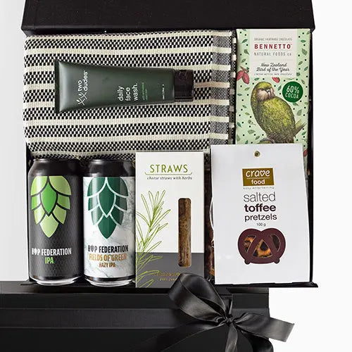 A luxury black gift box with magnetic closure containing a variety of gift items including a Turkish hand/gym towel, men's daily face wash, two cans of craft beer, a box of herb and cheese straws, and a salted toffee pretzel snack, with a keepsake gift box and free gift card visible.