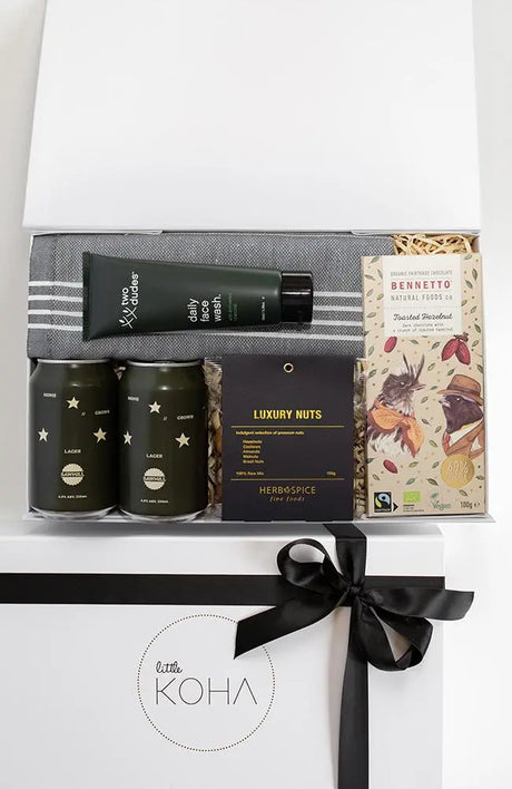 A gift box present tied with a black ribbon, containing various items including two cans of beer, a luxury nut mix, organic chocolate, a Turkish hand towel, and a daily face wash.