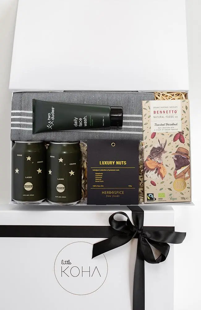 A gift box present tied with a black ribbon, containing various items including two cans of beer, a luxury nut mix, organic chocolate, a Turkish hand towel, and a daily face wash.