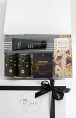 A gift box present tied with a black ribbon, containing various items including two cans of beer, a luxury nut mix, organic chocolate, a Turkish hand towel, and a daily face wash.